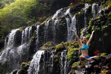 Heyecanlı Kafkas kadın kaya üzerinde oturma, şelale önünde silah yükselterek. Banyu wana amertha şelalesi, Wanagiri, Bali, Endonezya