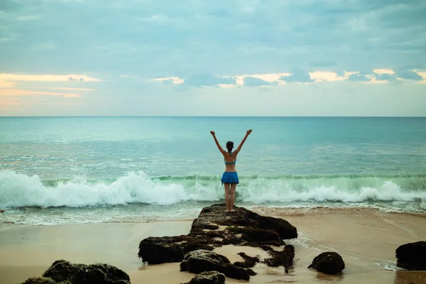 Giovane donna eccitata che alza le braccia sulla spiaggia di fronte all'oceano. Vista da dietro. Tramonto in spiaggia. Bali, Indonesia . — Foto Stock