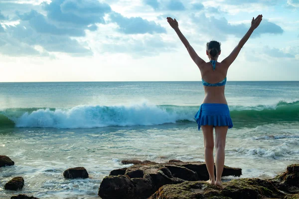 Giovane donna eccitata in piedi sulla roccia, alzando le braccia sulla spiaggia di fronte all'oceano. Vista da dietro. Tramonto in spiaggia. Viaggio di lusso in Asia. Onde oceaniche. Ricevuto. Tegal Wangi, Bali — Foto Stock