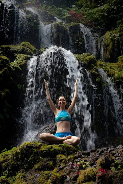 Heyecanlı Beyaz kadın kayanın üzerinde oturuyor ve şelalenin önünde kollarını kaldırıyor. Banyu Wana Amertha şelale Wanagiri, Bali, Endonezya. 