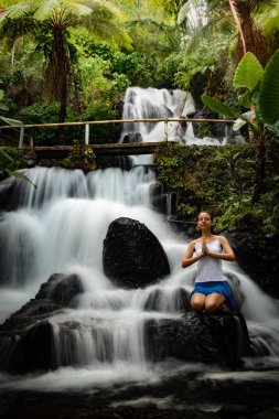 Genç Beyaz kadın meditasyon yapıyor, şelalede yoga yapıyor. Namaste mudra. Yavaş deklanşör hızı, hareket fotoğrafçılığı. Jembong şelalesi. Bali, Endonezya