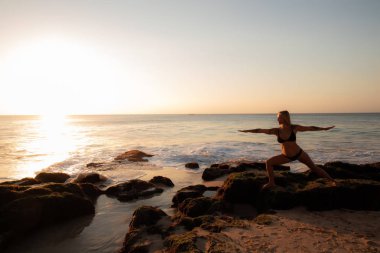 Yoga yapan genç kadın, Virabhadrasana'da sahilde poz veriyor. Gün batımı zamanı. Tegal Wangi plajı, Bali