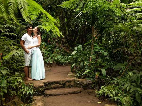 Щаслива усміхнена багаторасова пара обіймає і цілує по дорозі тропічним лісом. Молоде подружжя змішаних рас у відпустці в Азії. Убуд, Балі, Індонезія. — стокове фото