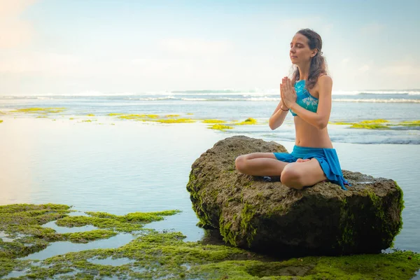 Young woman sitting on the rock, meditating, practicing yoga and pranayama at the beach, Bali. Hands in namaste mudra