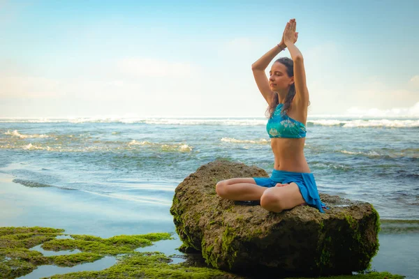 Young woman sitting on the rock, meditating, practicing yoga and pranayama at the beach, Bali. Hands raising in namaste mudra
