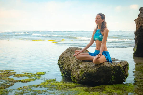 Young woman sitting on the rock, meditating, practicing yoga and pranayama at the beach, Bali. Hands in gyan mudra