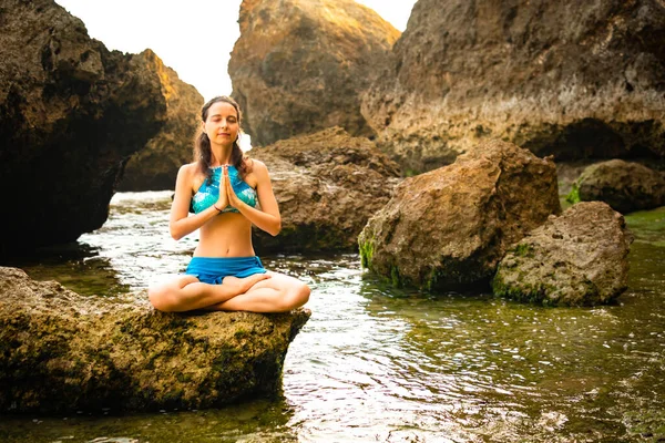 Молодая женщина сидит на скале, медитирует, практикует йогу и пранаяму на пляже, Бали. Руки в мудре намасте — стоковое фото