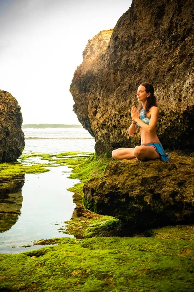 Young woman sitting on the rock, meditating, practicing yoga and pranayama at the beach, Bali. Hands in namaste mudra