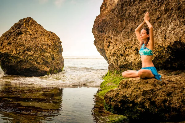 Young woman seating on the rock, meditating, practicing yoga and pranayama at the beach, Bali. Hands raising in namaste mudra.