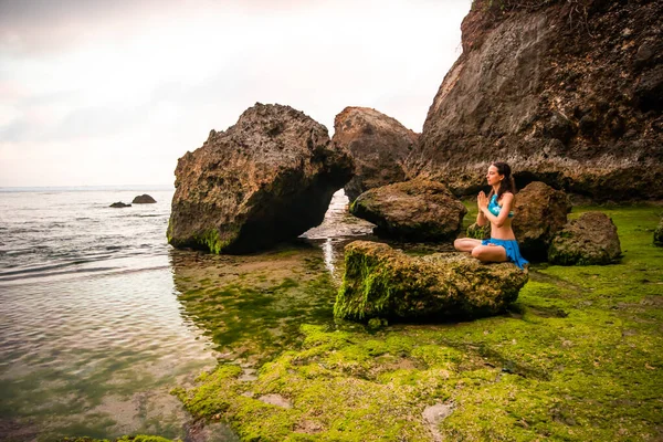 Mujer meditando, practicando yoga y pranayama. Namaste mudra. Postura de loto. Rocas cubiertas por algas verdes. Retiro de yoga, Bali, Playa Nyang Nyang —  Fotos de Stock