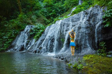 Kafkas kadın kayanın üzerinde duruyor, yoga yapıyor. Şelale yakınlarında namaste Mudra ile kol kaldıran genç bir kadın. Yoga dışarıda. Pucak Manik şelalesi Wanagiri, Bali, Endonezya. Arkadan görüntüle.