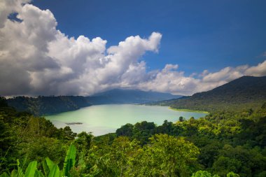 Panoramik manzara. Tepeler, dağlar ve tropikal yağmur ormanlarıyla çevrili göl manzaralı. Beyaz bulutlu mavi gökyüzü. Parlak güneş ışığı. Bedugul 'daki Buyan Gölü, Bali, Endonezya