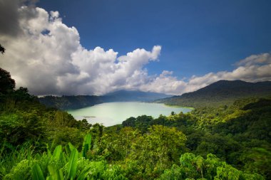 Panoramik manzara. Tepeler, dağlar ve tropikal yağmur ormanlarıyla çevrili göl manzaralı. Beyaz bulutlu mavi gökyüzü. Parlak güneş ışığı. Bedugul 'daki Buyan Gölü, Bali, Endonezya