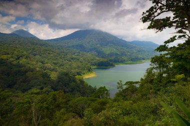 Panoramik manzara. Tepeler, dağlar ve tropikal yağmur ormanlarıyla çevrili göl manzaralı. Beyaz bulutlu mavi gökyüzü. Parlak güneş ışığı. Bedugul 'daki Buyan Gölü, Bali, Endonezya