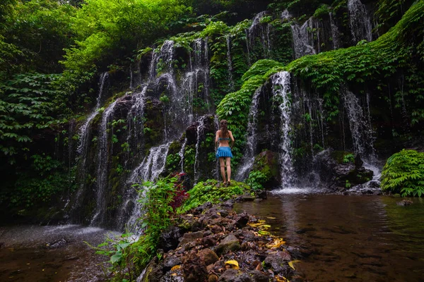 Young traveler woman at waterfall in tropical forest. Caucasian woman wearing blue bikini. View from back. Travel concept. Banyu Wana Amertha waterfall in Bali. Slow shutter speed, motion photography