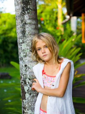 Ağacın yanında duran, beyaz elbiseli ve pembe yüzme takımlı şirin küçük kız. Dalgın duygularla yüzleş. Tropikal bahçede vakit geçirmek. Çocukluk konsepti. Bali, Endonezya