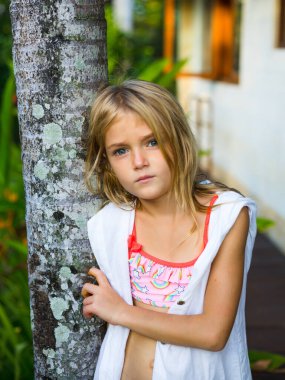 Ağacın yanında duran, beyaz elbiseli ve pembe yüzme takımlı sevimli küçük kız. Dalgın duygularla yüzleş. Tropikal bahçede vakit geçirmek. Çocukluk konsepti. Bali, Endonezya