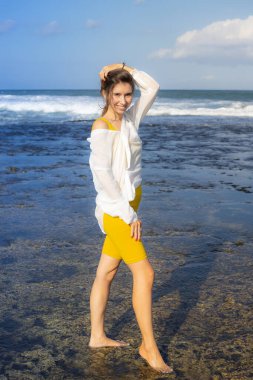 Dalgalar çekildiğinde sahilde yürüyen güzel genç bir kadın. Beyaz bluz ve sarı spor giysili beyaz kadın. Mutluluk ve özgürlük. Seyahat yaşam tarzı. Uzayı kopyala Bali, Endonezya