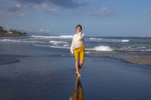 スリムな若い女性は黒砂のビーチを歩く 黄色のスポーツウェアと白いブラウスを着た白人女性 幸せと自由 旅行生活 コピースペース インドネシアのバリ — ストック写真