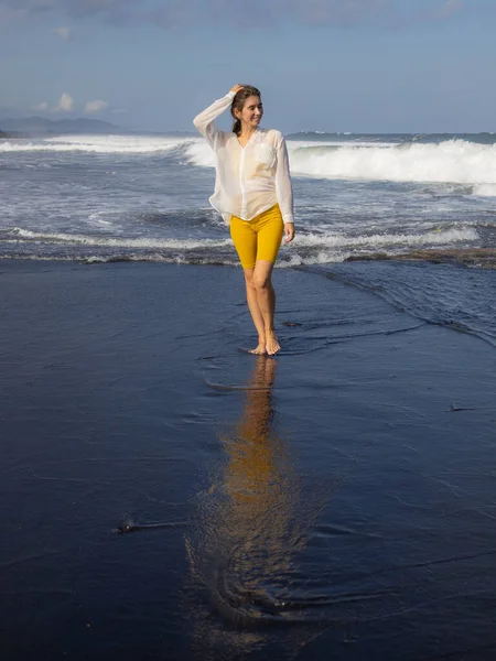 Beautiful young woman walking on black sand beach. Caucasian woman wearing yellow sportswear and white blouse. Happiness and freedom. Travel lifestyle. Copy space. Bali, Indonesia