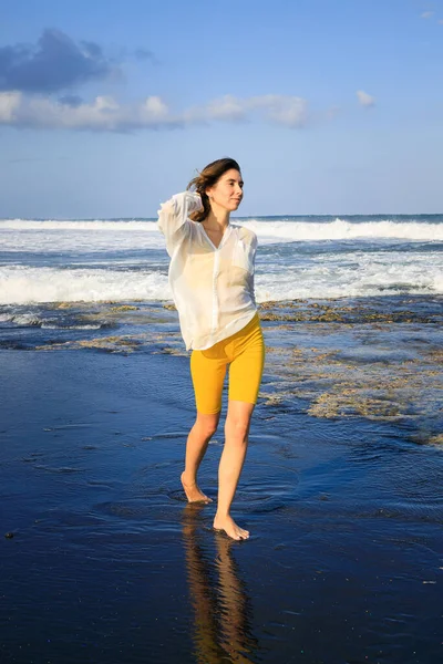 若い女性が黒い砂浜を歩いている 黄色のスポーツウェアと白いブラウスを着た白人女性 幸せと自由 旅行生活 コピースペース インドネシアのバリ — ストック写真
