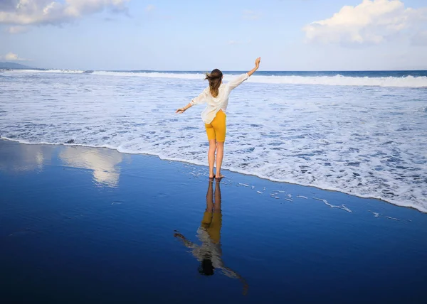 美しい若い女性が黒い砂のビーチを歩いている 黄色のスポーツウェアと白いブラウスを着た白人女性 水の反射 旅行生活 後ろから見るんだ コピースペース インドネシアのバリ — ストック写真