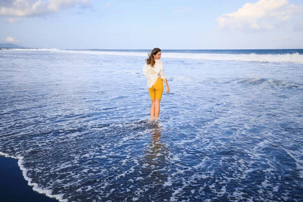 美しい若い女性が黒い砂のビーチを歩いている 黄色のスポーツウェアと白いブラウスを着た白人女性 幸せと自由 旅行生活 コピースペース インドネシアのバリ — ストック写真