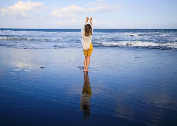 美しい若い女性が黒い砂のビーチを歩いている 黄色のスポーツウェアと白いブラウスを着た白人女性 水の反射 旅行生活 後ろから見るんだ コピースペース インドネシアのバリ — ストック写真
