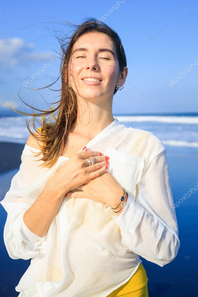 Portrait of beautiful smiling woman with jewelry. Closed eyes. Hair and clothes are blown by the wind. Happy Caucasian woman enjoying sunlight. Travel lifestyle. Bali, Indonesia