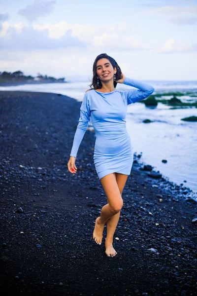 Beautiful young woman walking on black sand beach. Caucasian woman wearing blue mini dress. Romantic concept. Travel lifestyle. Bali, Indonesia