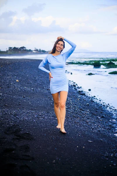 Siyah kumsalda yürüyen güzel genç bir kadın. Mavi mini elbise giyen beyaz bir kadın. Romantik bir kavram. Seyahat yaşam tarzı. Bali, Endonezya