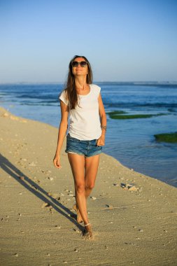 Sıska genç bir kadın sahilde yalınayak yürüyor. Beyaz kadın kot şort giyiyor, beyaz tişört giyiyor, güneş gözlüğü takıyor. Mutluluk ve özgürlük. Seyahat yaşam tarzı. Açık hava konsepti. Uzayı kopyala Bali