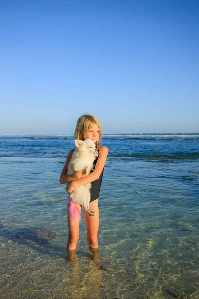 可爱的小女孩在海滩上抱着一条粉红尾巴的小白花狗 童年的概念 穿着黑色游泳衣的金发女孩 花时间在户外 低潮时的海洋 — 图库照片