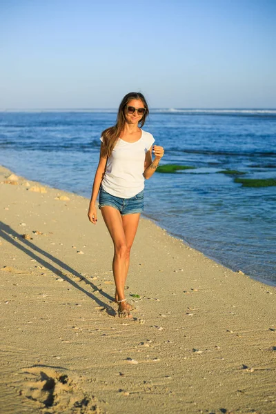 スリムな若い女性がビーチに沿って裸足で歩く ジーンズのショートパンツ 白いTシャツ サングラスをかけている白人女性 幸せと自由 旅行生活 屋外のコンセプト コピースペース バリ島 — ストック写真