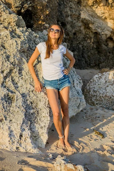 スリムな若い女性が岩の近くの砂の上に裸足で立っている ジーンズのショートパンツ 白いTシャツ サングラスをかけている白人女性 ビーチで日光を楽しむ アジアでの休暇 旅行のコンセプト バリ島 — ストック写真