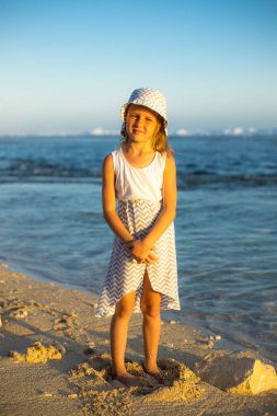 Kumsalda tatlı küçük bir kız. Kafkasyalı bir kız, elbise ve şapka takıyor, okyanus kıyısındaki kumun üzerinde duruyor. Mutlu bir çocukluk. Yaz kavramı. Çocukla yolculuk. Melasti plajı, Bali, Endonezya 