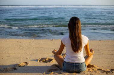 Genç bir kadın nilüfer çiçeğiyle kumlara oturmuş okyanusun önünde poz veriyor. Sahilde yoga. Eller Gyan Mudra 'da. Meditasyon konsepti. Arkadan bak. Uzayı kopyala Melasti plajı, Bali, Endonezya
