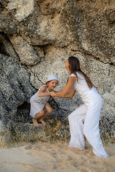 Anne kızıyla kayaların yanındaki sahilde oynuyor. Aile kavramı. Kız atlaması. Anne ve kızı arasındaki ilişki. Dışarıda vakit geçir. Mutlu bir çocukluk. Melasti plajı, Bali, Endonezya