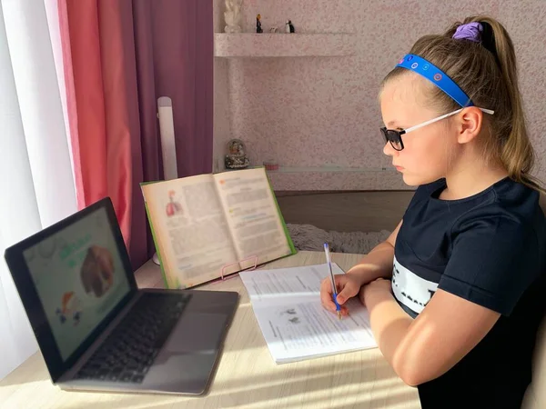 The girl at the table, writes tests. The schoolgirl is engaged, remotely on the laptop. School accessories on a table, for the pupil.