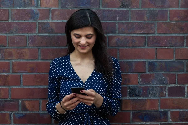 Tuğla duvara yaslanmış ise telefon kullanarak gülümseyen genç kadın