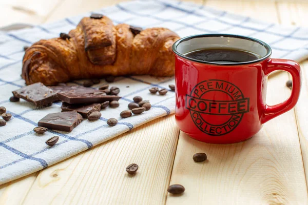 Red coffee mug and croissant on wooden background. The view from the top. Coffee beans and chocolate