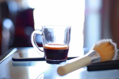 Bir fincan kahve, telefon, makyaj seti ve cam sehpada fırçalar.