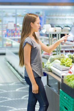  Genç kız süpermarkette sebze reyonunda alışveriş yapıyor. Ürün bilgileri okumak, günlük ürünleri seçmek. Gençlere göre bilinçli sağlıklı yiyecek seçimi kavramı.