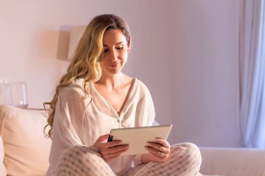Orta yaşlı zarif bir kadın kanepede oturuyor ve tabletten haberler okuyor. Evde tablet pc kullanılıyor.
