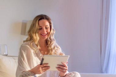 Orta yaşlı zarif bir kadın kanepede oturuyor ve tabletten haberler okuyor. Evde tablet pc kullanılıyor.