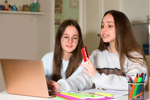 Two teen girls surf the Internet on their laptop, look for products of interest to them and make purchases with a credit card. Teenagers\' concept of navigation, research and shopping
