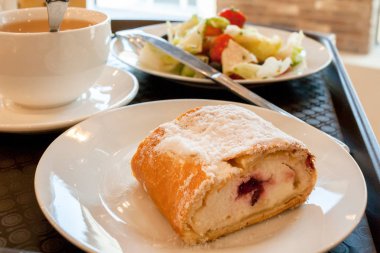 Beyaz tabaklarda: kiraz strudel, yunan salatası ve kahverengi salver üzerinde bir fincan çay.