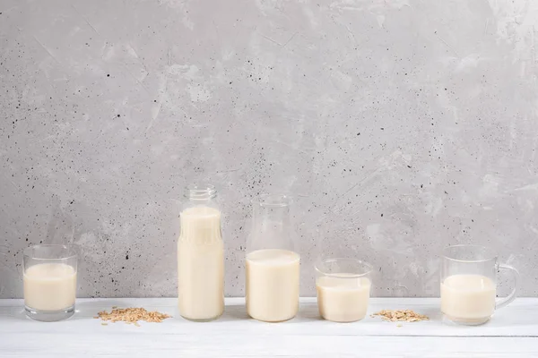 Set of glass containers with oat milk on white wooden table on concrete wall background.