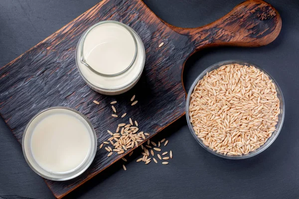 Dairy free oat milk in glass bottle and glass and oat seeds in glass bowl on wooden kitchen board on grey background.