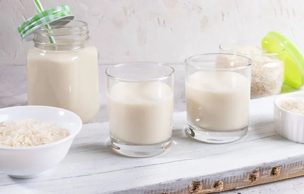 Bicchieri, tazza di vetro in muratore con latte di riso, ciotola e vaso con semi rialzati su tavola di legno su sfondo bianco . — Foto Stock